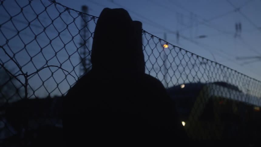 A silhouetted figure in a hoodie watches a train pass by from behind a chain-link fence at dusk