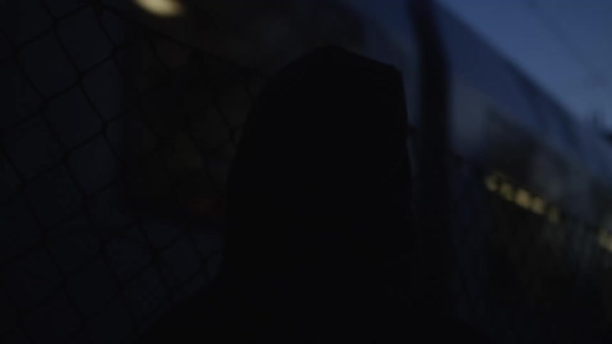 A mysterious silhouette of a person in a hoodie stands by a chain-link fence, watching a train pass by in the dark