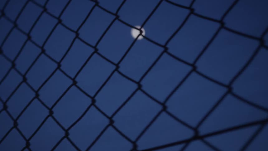 A blurry view of the moon in the night sky is seen through a dark, out-of-focus chain-link fence
