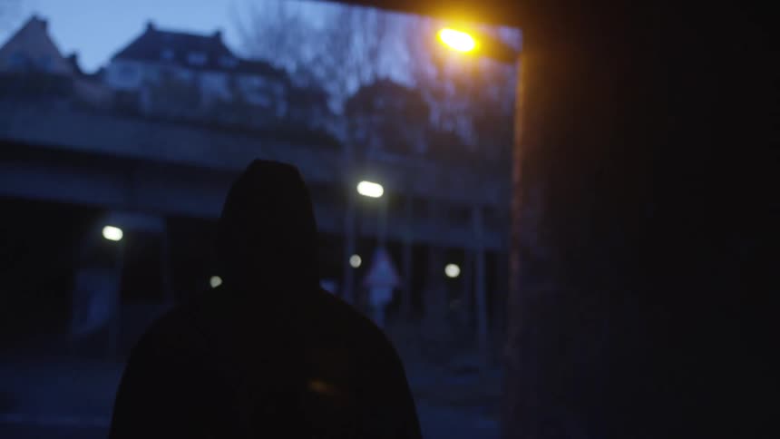 A mysterious hooded figure walks in silhouette through a dark urban underpass at night