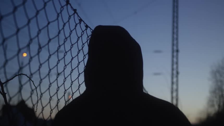 The silhouette of a mysterious person wearing a hoodie stands still next to a chain-link fence at dusk