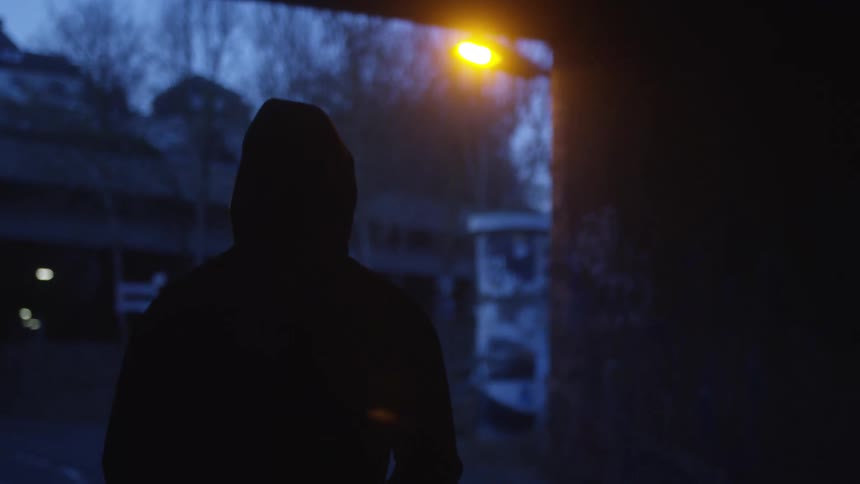 A mysterious silhouetted figure in a hoodie stands in a dark urban underpass at night