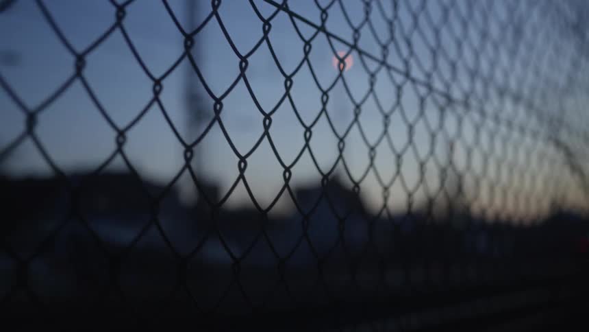 A chain-link fence is silhouetted against the dim light of dusk in an urban setting