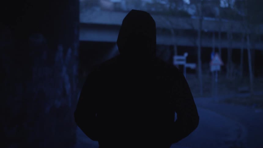 Slow motion shot of a mysterious silhouetted figure in a hoodie walking out of a dark, graffiti-covered tunnel at night