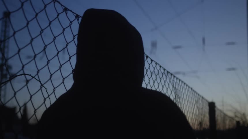 A silhouetted person in a hoodie contemplates from behind a chain-link fence as night falls on an industrial landscape