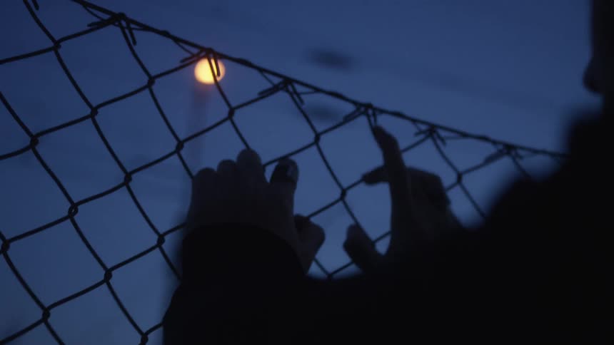 A silhouetted person's hands clutch a chain-link fence at dusk, conveying a sense of loneliness and despair