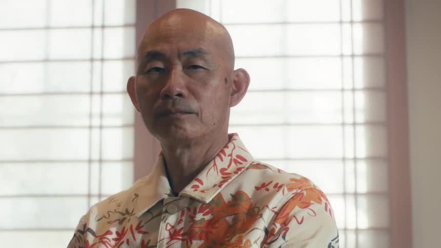 An intense and stoic portrait of an elderly bald Asian man staring into the camera as it slowly zooms in