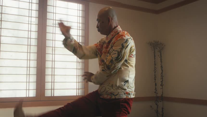 An elderly Asian man gracefully practices a slow martial arts routine in a serene room with Japanese-style windows