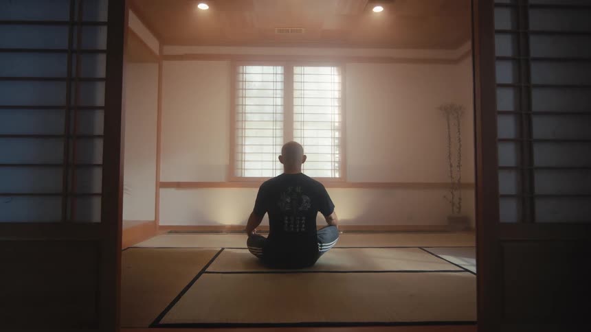 A man sits in silent meditation on a tatami mat floor in a traditional Japanese-style room, facing a bright, hazy window