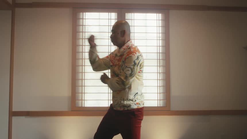 An elderly Asian man in a colorful shirt gracefully practices Tai Chi in a serene room with a shoji screen window