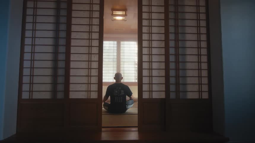 Traditional Japanese shoji screen doors open to reveal a man meditating alone in a serene, sunlit room