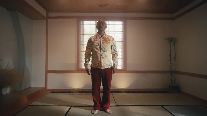A barefoot man in a colorful jacket stands perfectly still in a traditional, minimalist Japanese room