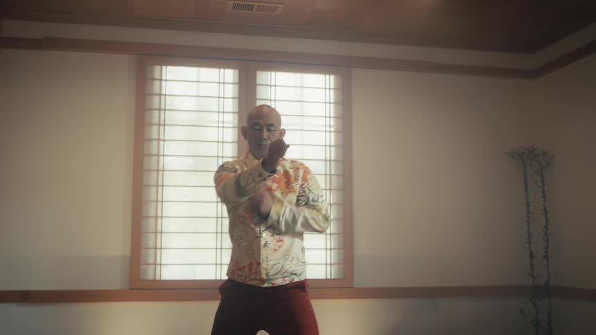 A bald Asian man gracefully practices slow, deliberate tai chi movements in a serene, minimalist dojo