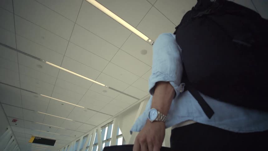 Low-angle follow shot of a man with a backpack and rolling suitcase walking through a modern airport corridor