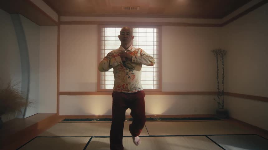 A bald man gracefully practices a slow Tai Chi or Qigong routine in a tranquil, traditional Japanese room as the camera slowly zooms out