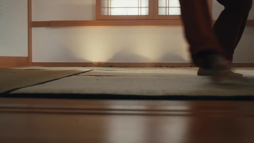 A low-angle close-up shows a person's bare feet gracefully practicing martial arts on a traditional tatami mat floor