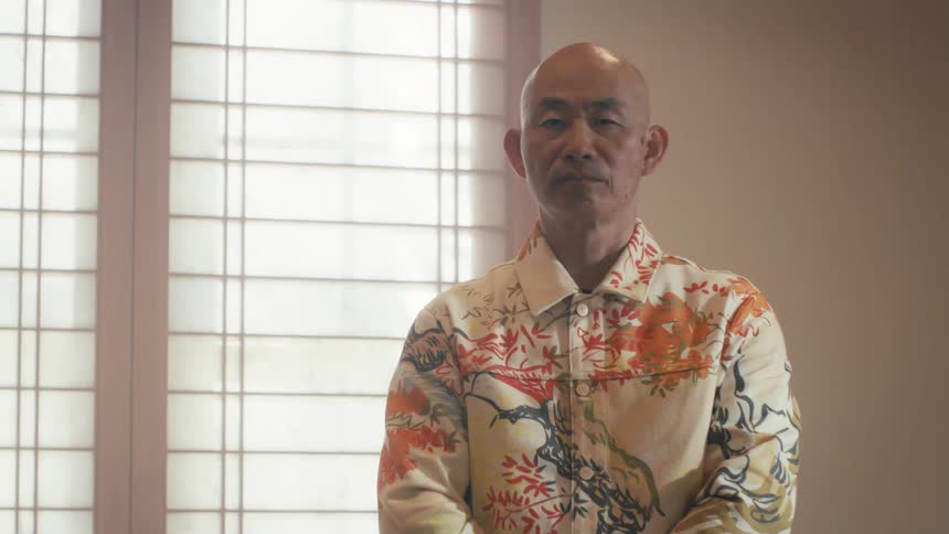 A serious older Asian man in a vibrant patterned shirt stands for a portrait in front of a traditional shoji screen