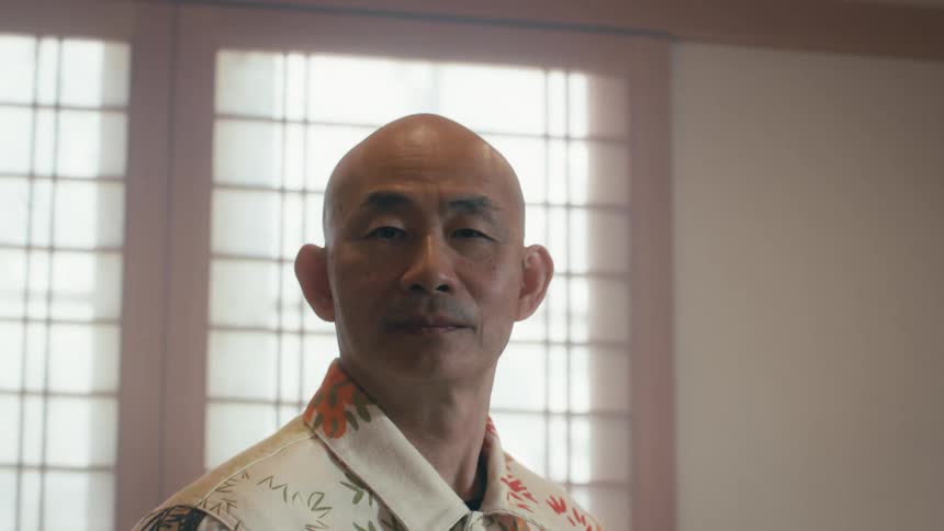 A blurry shot of an older man comes into focus as he stands in a soft-lit room with a serious expression