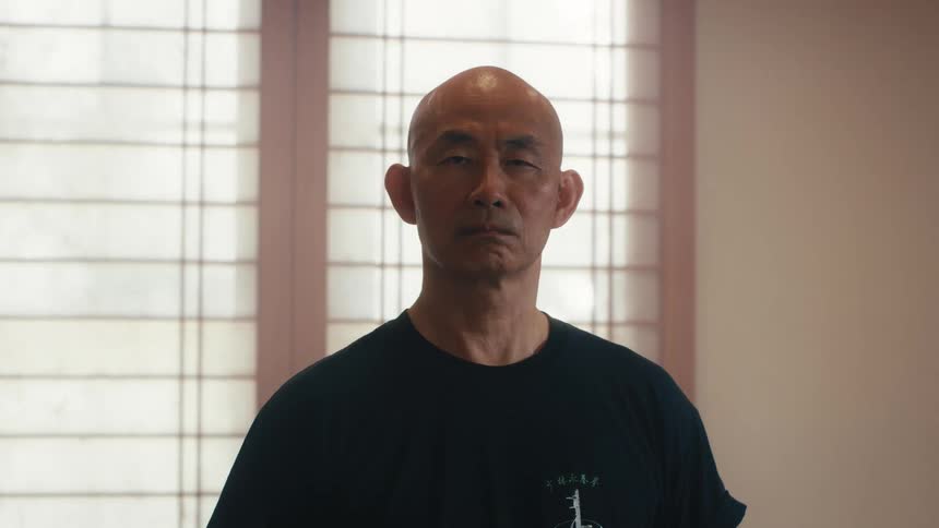 Stern portrait of a bald, senior Asian man staring intently at the camera in front of a shoji screen window