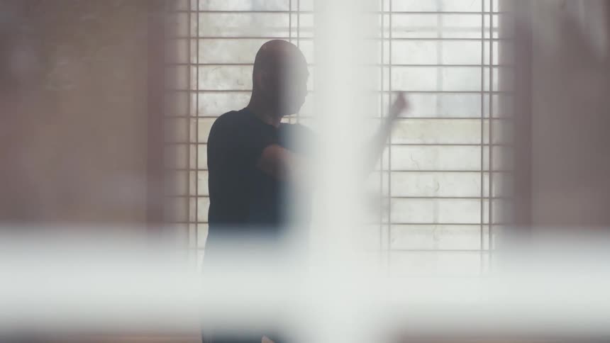 A focused bald man practices the slow and deliberate movements of martial arts in a softly lit dojo with a traditional Japanese screen