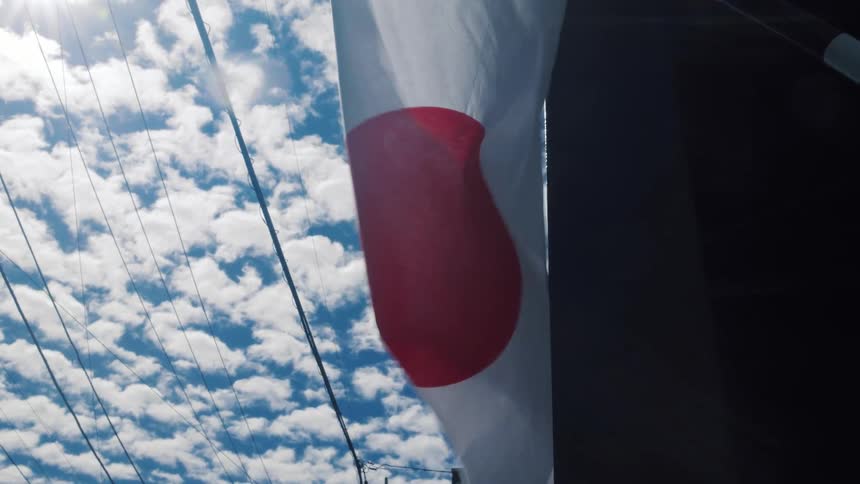 A low angle shot of the Japanese flag waving in the wind against a blue sky with fluffy clouds and sun flare