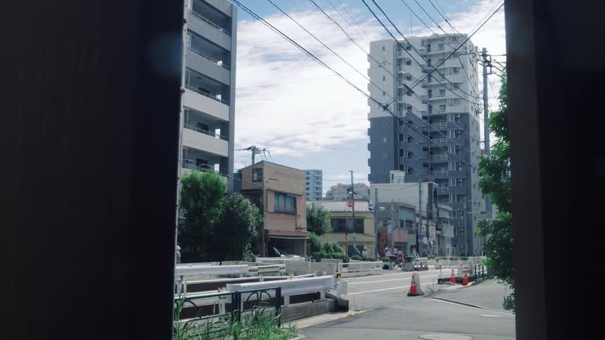 A slow zoom-out from a shaded viewpoint reveals a quiet street in a Japanese city with modern buildings and power lines on a bright day