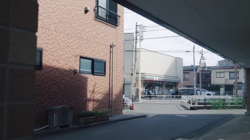 A quiet alleyway in Japan looks out onto a street with a convenience store and pedestrians