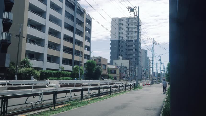 An elderly man walks down a quiet sidewalk lined with modern apartment buildings in a Japanese city