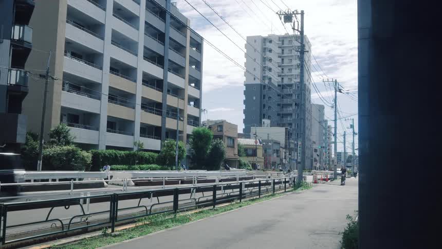 A quiet residential street in a Japanese city shows cars passing by large apartment buildings on a bright day