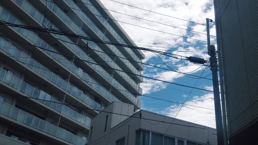 A static low-angle shot captures a modern apartment building and a web of power lines against the cloudy blue sky in a Japanese city