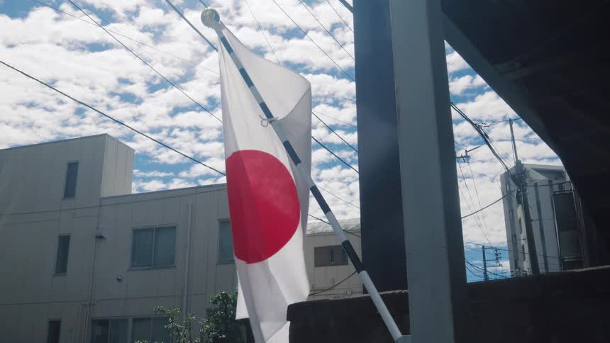 A slow-motion, low-angle shot of the Japanese flag waving in an urban setting against a blue sky with clouds
