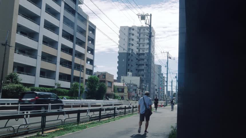 An elderly man walks down a sunny sidewalk alongside traffic in a Japanese city