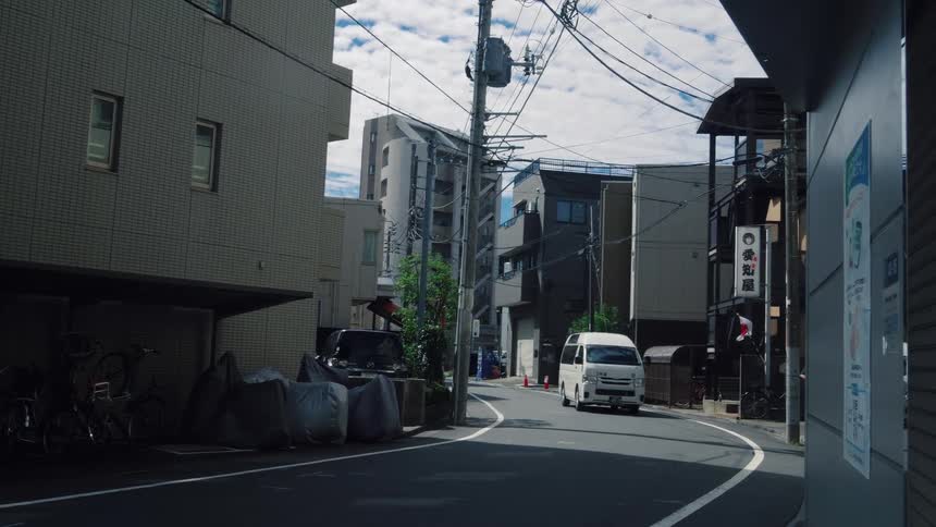 A white passenger van drives slowly down a quiet, curved residential street in a Japanese city during the day