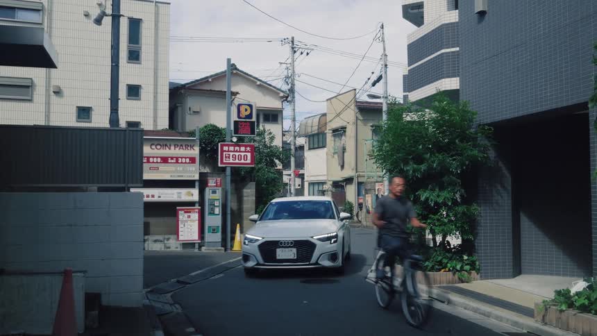 A white sedan navigates a narrow city street in Japan as a man rides by on his bicycle