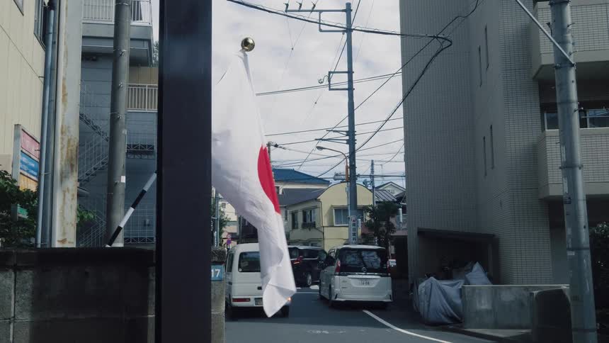 A Japanese flag gently waves in the foreground of a quiet residential street in Tokyo as cars drive by