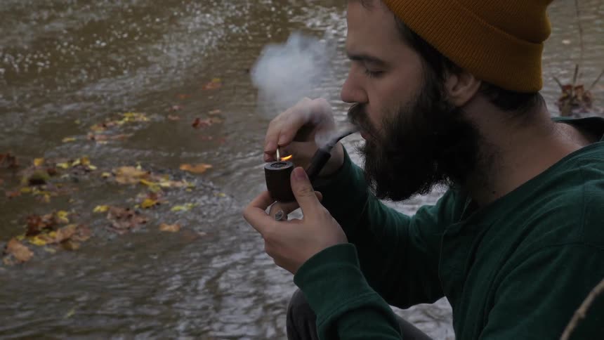 Man lights a pipe with a match next to a stream