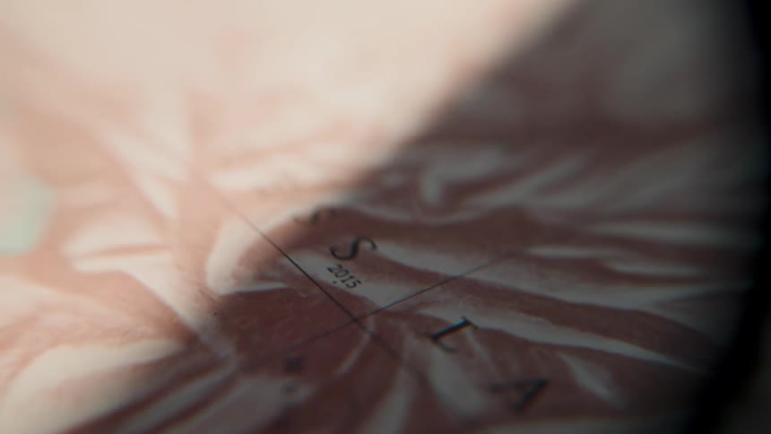 A moody, extreme close-up shot shows a shadow moving slowly across a vintage, sepia-toned map