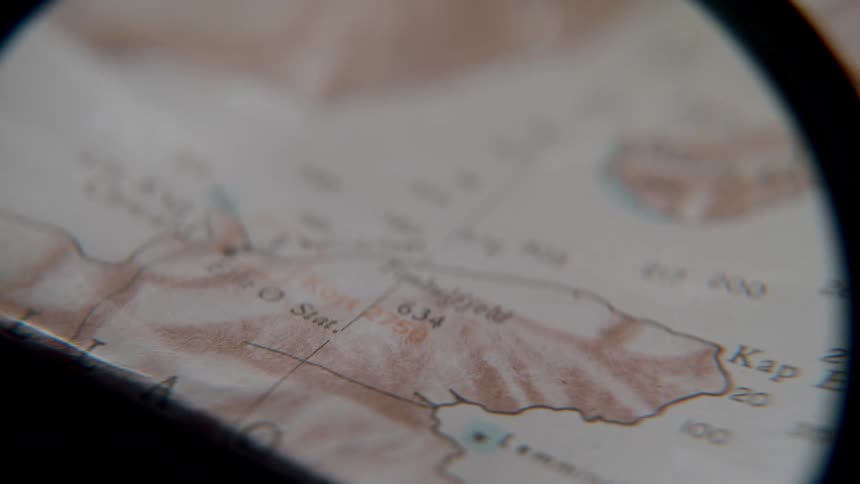 A magnifying glass slowly moves over a vintage topographical map, searching for a specific location or route