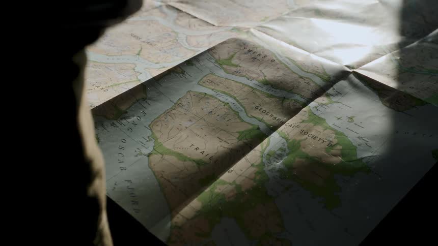 A light source moves across a vintage map of Greenland's fjords as a silhouetted explorer plans an expedition
