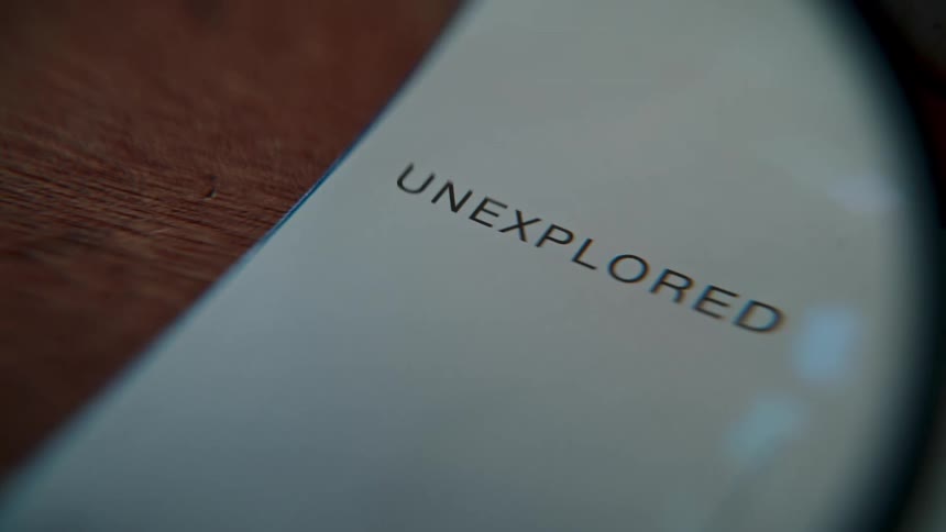 A close-up rack focus shot reveals the word 'unexplored' printed on a piece of paper sitting on a wooden table