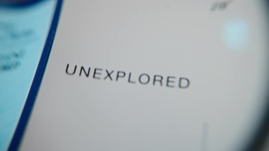 A magnifying glass slowly focuses on the word 'Unexplored' printed on a map, invoking a sense of mystery and adventure