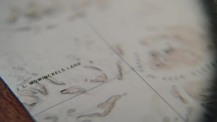 A close-up, handheld shot of a vintage topographical map focusing on the name Mowinckels Land