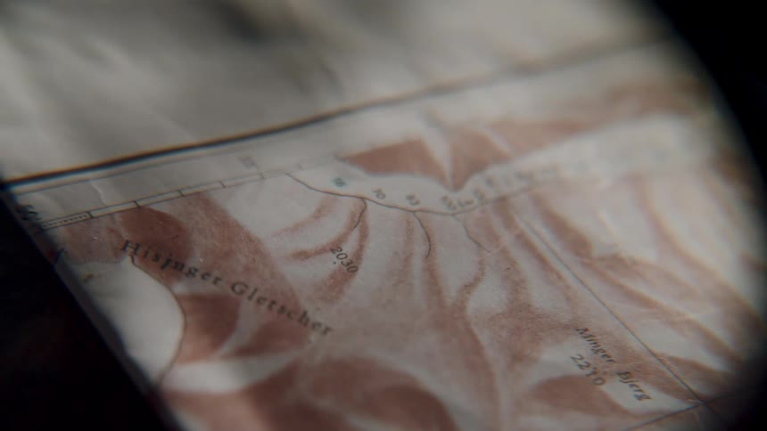 A slow, moody pan across a vintage topographical map reveals the details of a glacier, suggesting an upcoming expedition or adventure