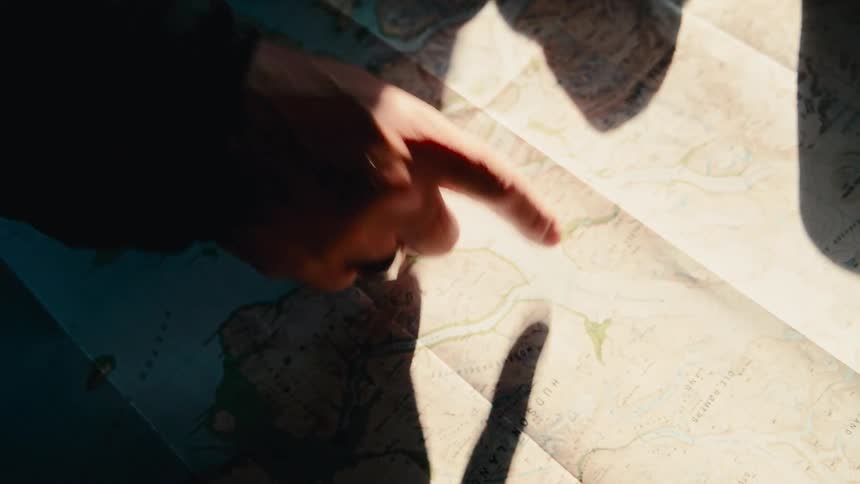 A person's finger traces a route on a topographical map, planning an adventurous journey in dramatic sunlight
