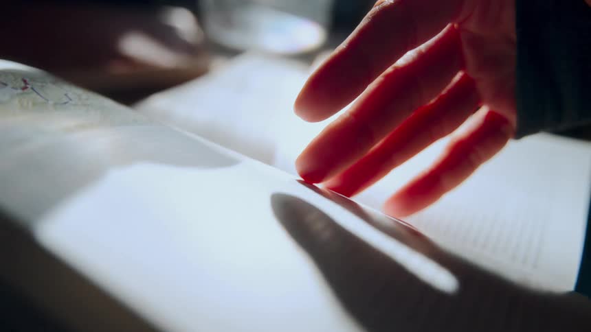 A hand gently turns the pages of an illustrated history book as sunlight dances across the text and images