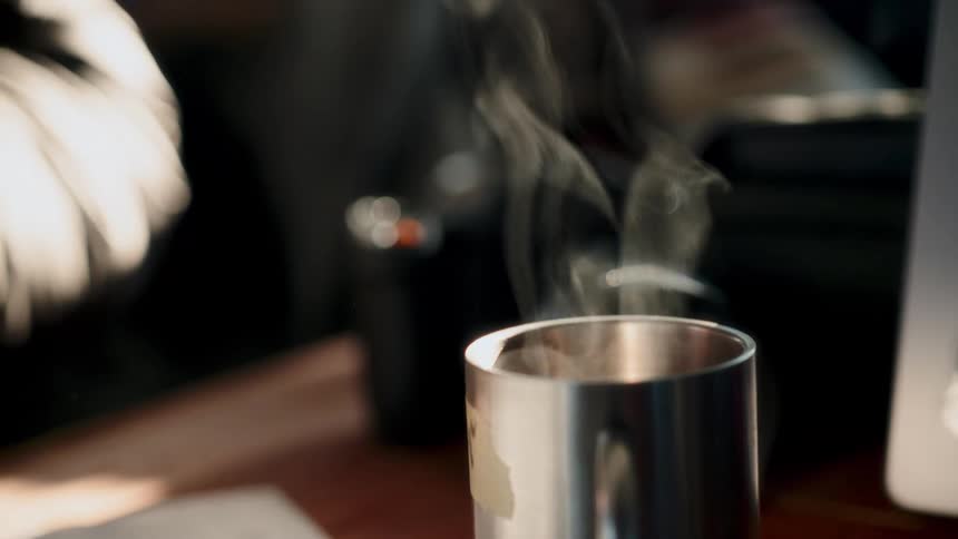 Steam rises in slow motion from a hot beverage in a stainless steel mug sitting on a sunlit desk