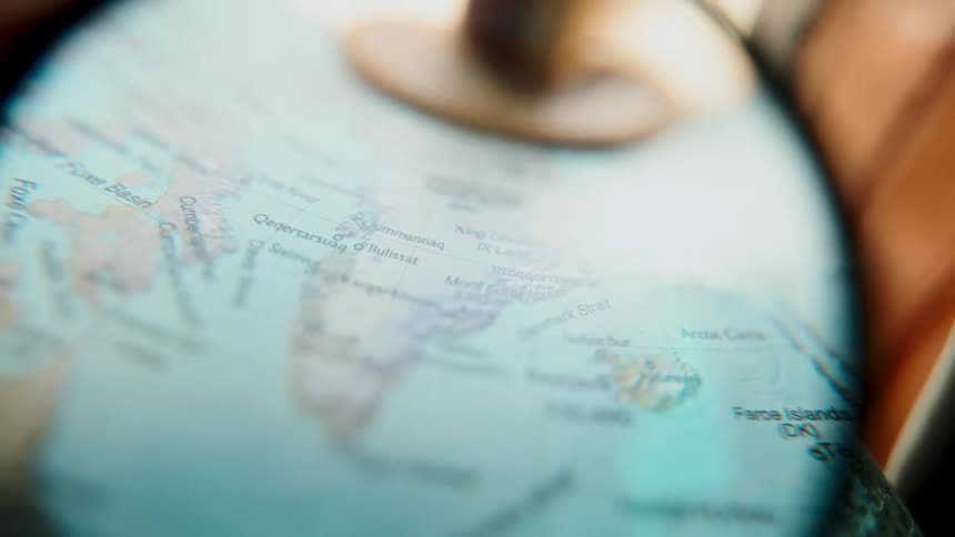 A person uses a magnifying glass to examine a world globe, focusing on Greenland and Iceland