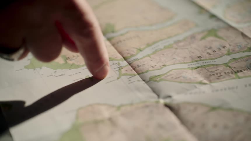 A close-up of a finger with a ring tracing a path along a fjord on a vintage paper map, with sunlight casting a strong shadow