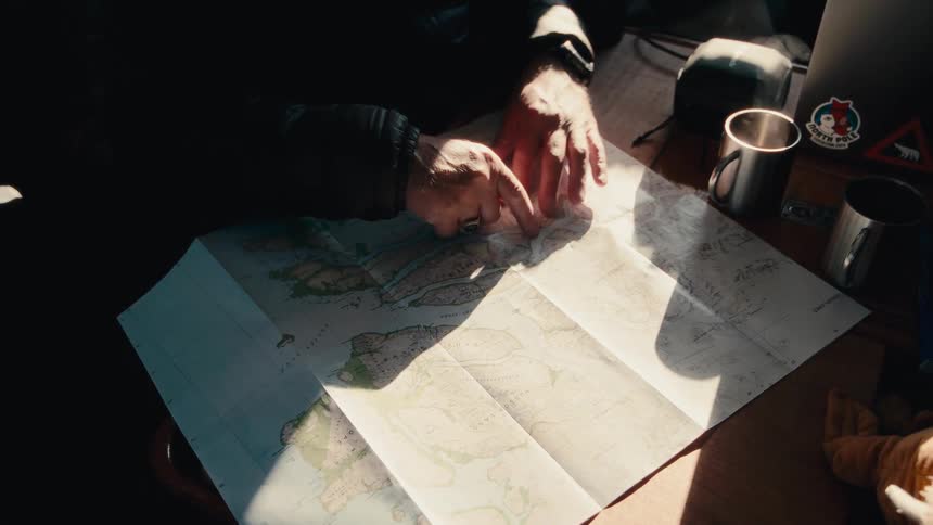 An adventurer traces their finger along a route on a paper map while planning their next expedition inside a sunlit room