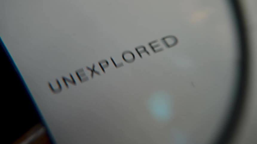 A macro close-up shot examines the word 'unexplored' printed on white paper through a magnifying glass